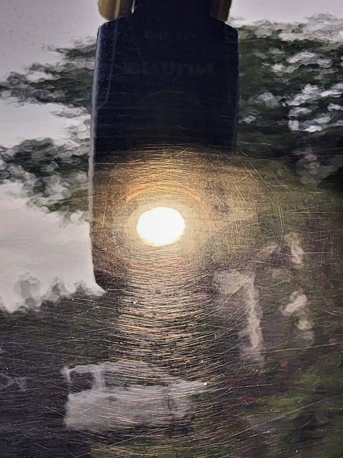 The image depicts a sun-like reflection in the center of a glossy surface, surrounded by a radial pattern of scratches. The reflection is bright and clear, and the scratches give it the appearance of a shining sun. In the background, there are inverted and distorted reflections of trees and the silhouette of a person standing, indicating the photo was taken on a reflective surface outdoors during the day.
