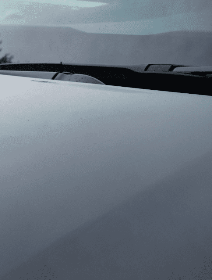 This is an image showing a close-up view of a car's hood and windshield with the wiper blades in rest position, under overcast lighting. The focus is on the smooth, shiny surface of the hood which reflects the grey, muted light of the sky.