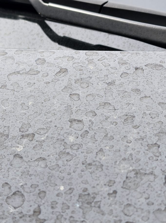 A detailed image of raindrops scattered across a grey textured surface, with a partial view of a car's black wiper blade in the upper left corner. The varying sizes and shapes of the water droplets create a random pattern on the surface.