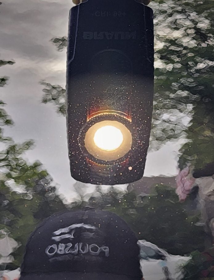 The image captures a reflective surface with a bright circular glare in the center, possibly from a light source like the sun. Above the glare, there's an inverted reflection of a cell phone or device with text, while below, a cap with a logo is visible, also reflected and inverted. The surface is speckled with water droplets, and there's a blurred background that includes trees, suggesting an outdoor setting.