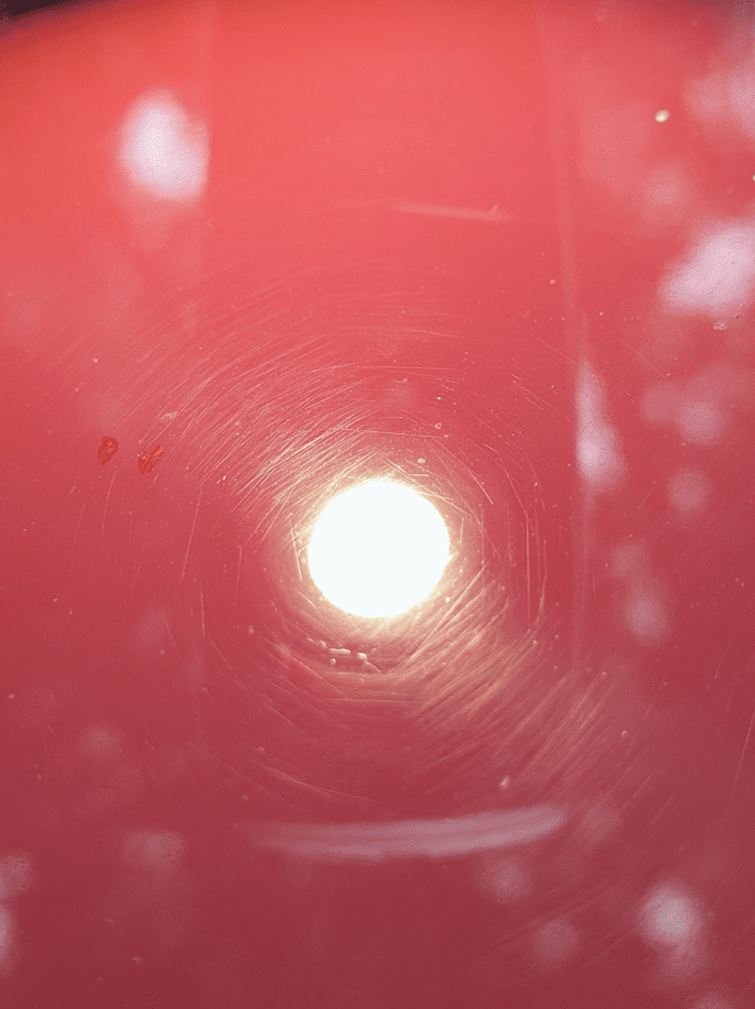 A close-up photograph showing a circular light reflection on a glossy red surface with visible concentric circular scratches, likely from polishing or cleaning. Small specks of dust are scattered around the light reflection.