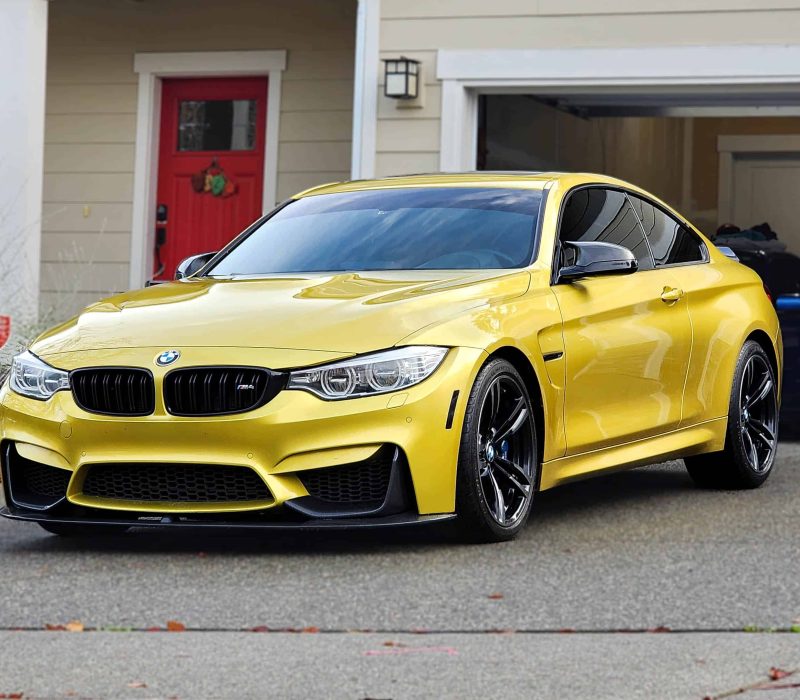 The image displays a bright yellow luxury sports coupe parked on a residential street. The car's design is dynamic and sleek, with a low profile, distinctive twin-kidney grille, and angular headlights, indicative of a high-performance model. A suburban home with a red door and an open garage revealing items stored inside serves as the backdrop, creating a contrast between the vivid car color and the neutral tones of the house.