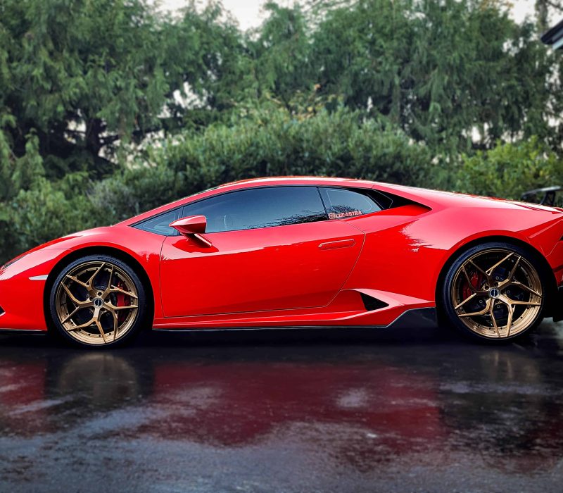 A striking red Lamborghini Huracán featuring gold rims, parked on a wet driveway with lush greenery in the background.