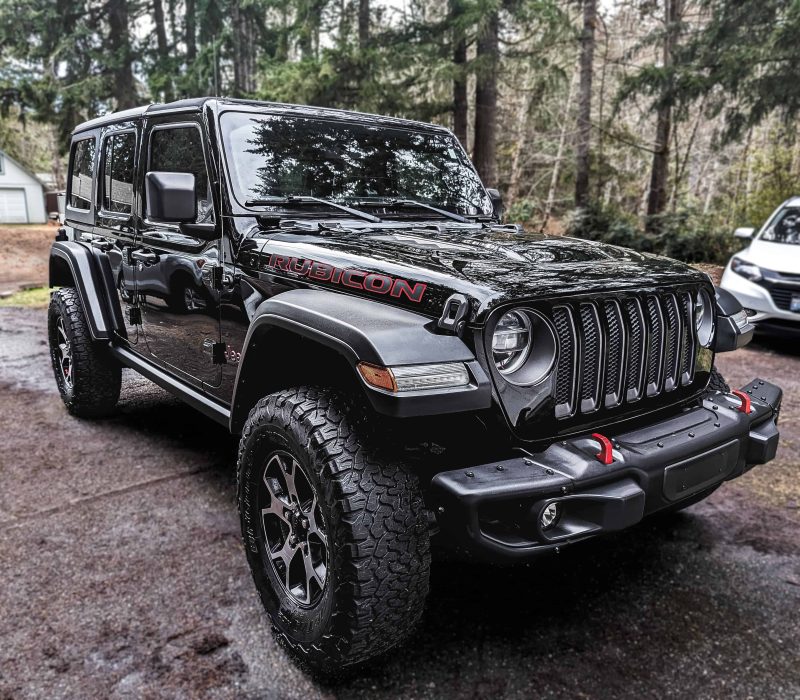 Black Jeep Wrangler Rubicon parked on a gravel driveway, showcasing its rugged design and off-road capabilities, with a forest in the background.